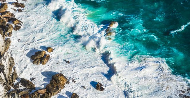 Aerial view of rough sea waves crashing on rocky coastline. The turquoise water is turbulent, with white foam being formed around the rocks. This image captures the dramatic and powerful nature of the ocean. Ideal for use in nature magazines, travel blogs, environmental campaigns, and educational resources on coastal environments.