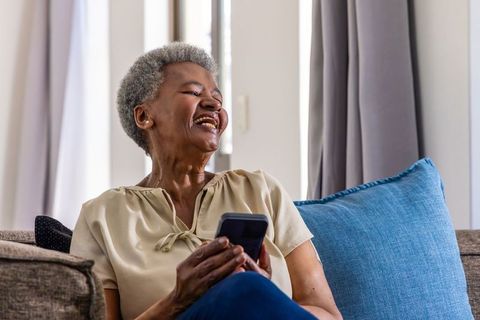 Senior african american woman laughing while holding smartphone on cozy living room sofa