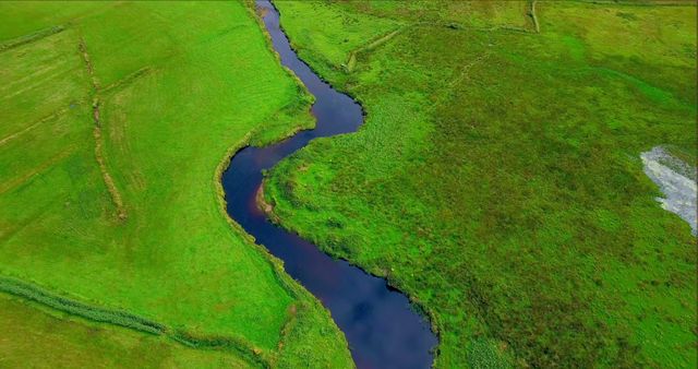 A winding river cuts through vibrant green fields in an aerial landscape view, showcasing the natural beauty of the meandering waterway. The lush greenery surrounding the river highlights the fertile nature of the land.