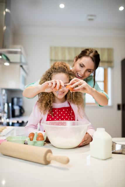 Mother assisting daughter in breaking eggs into a mixing bowl in a bright, modern kitchen. This image captures the essence of family bonding and the joy of cooking together. Ideal for use in articles or advertisements about family activities, cooking tutorials, parenting, and home life.