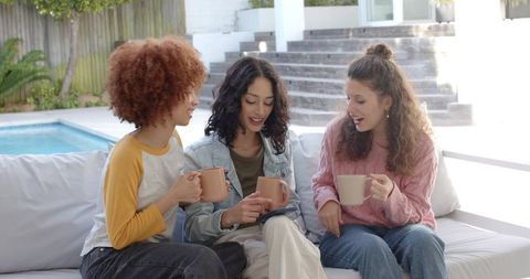 Diverse friends enjoying coffee and smartphone by sunlit poolside sofa