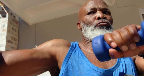 Mature man lifting dumbbell on sunlit porch wearing blue tank top, senior fitness strength