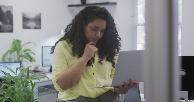 Businesswoman with curly hair stands in bright office, holding laptop and thinking. Ideal for images related to workplace productivity, strategic planning, businesswomen in technology, and modern office environments.