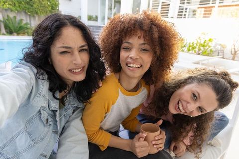 Multicultural friends smiling and taking selfie by backyard pool while holding mug