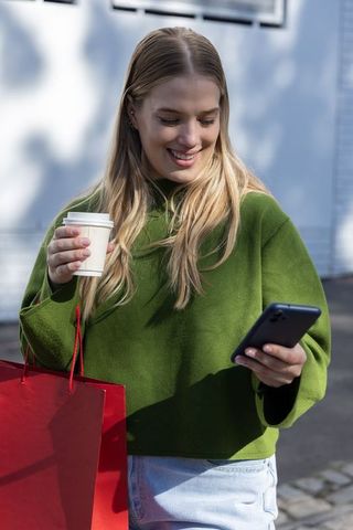 Smiling woman checking phone while holding coffee cup and red shopping bag on sunny street
