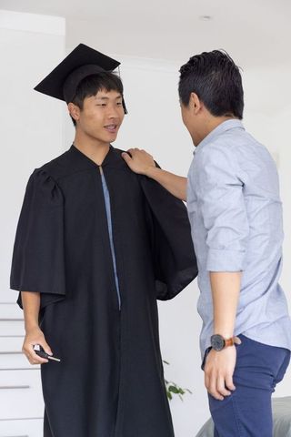Asian graduate wearing mortarboard and gown receiving supportive pat from friend at home
