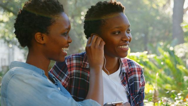 This captures joyful twin sisters sharing earphones while listening to music outside, symbolizing unity and companionship. Ideal for promoting themes of friendship, family bonding, and joyful leisure activities. Perfect for advertisements or articles focusing on music enjoyment and personal technology in everyday life settings.