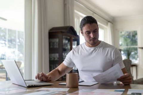 Remote work at home office: man reviewing paperwork with laptop and coffee at dining table