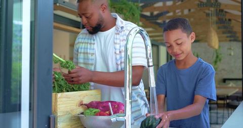 African american father and teen son washing fresh vegetables at modern kitchen sink