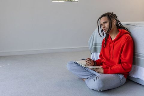 African american man writing in notebook while sitting on bedroom floor in natural light