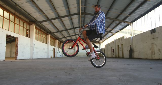 Man performing wheelie on a bicycle inside an empty warehouse with large windows and high ceiling. Ideal for use in advertisements focused on urban lifestyle, sports, physical activity, recreational cycling, and sports attire promotions.