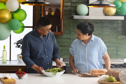 Mature couple preparing casual party meal in modern kitchen with balloons and fresh salad