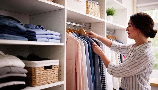Woman sorting shirts in organized closet with folded clothes, wicker baskets, natural light