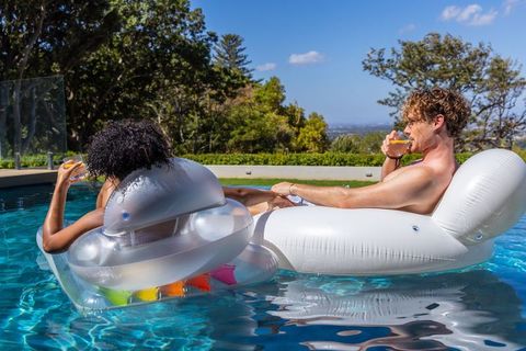 Sunlit pool lounging by mixed-race couple on clear white inflatables sipping orange drinks