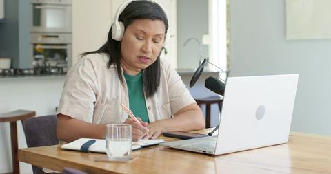 Mature woman taking notes during home podcast with laptop, headphones and microphone