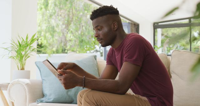 Image of relaxed african american man sitting on sofa and using tablet. relaxing and spending time with technology at home.