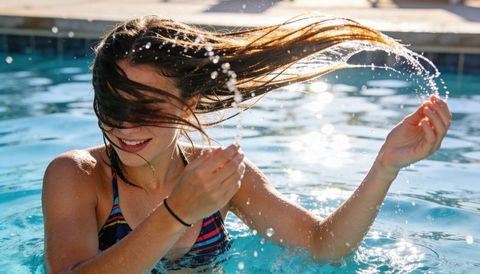 Young woman flipping hair back in sunlit backyard pool creating water arc and droplets
