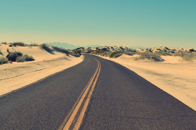 Picture showcases an empty desert road leading towards the horizon surrounded by sandy dunes under clear sky. Suitable for themes related to travel, adventure, road trips, solitude, and natural landscapes. It can be used for websites, travel blogs, promotional materials, or background images for inspirational quotes.