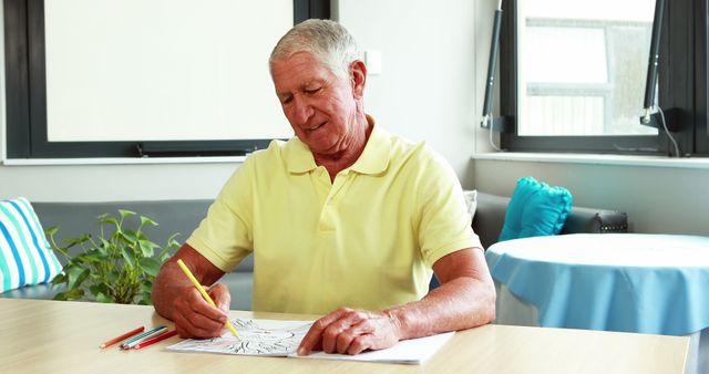 Senior man engaging in a relaxing art therapy activity, focusing on coloring an ornate design while sitting at a table in a well-lit room. This image can be used to promote mental health benefits for elderly people, art therapy sessions, or senior activities. Suitable for use in articles and blogs about wellness, hobbies for seniors, and creative therapies for mental health.
