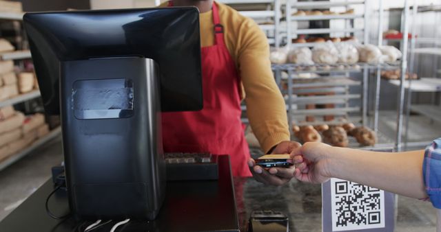 Customer making a contactless payment using a smartphone at a bakery counter. Cashier wearing a red apron processes the transaction. Bakery shelves with various bread and bagels in the background. Ideal for topics related to modern payment methods, retail technology, and small business operations.