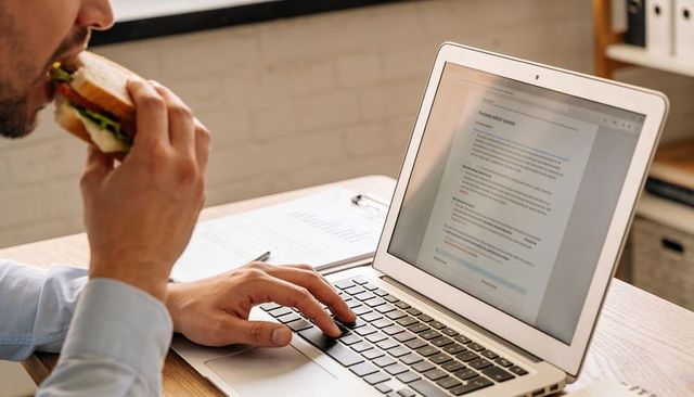 Eating lunch while working, man eating sandwich typing on laptop at office desk