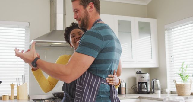 Image of happy diverse couple dancing in kitchen in aprons. Love, relationship and spending quality time together at home.