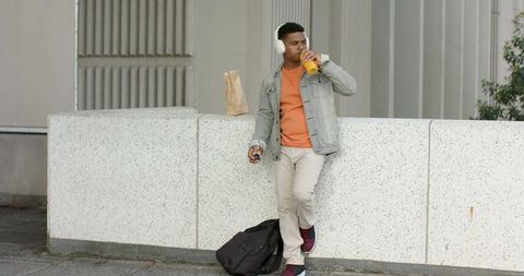 African american man leaning on concrete wall drinking juice wearing headphones carrying backpack