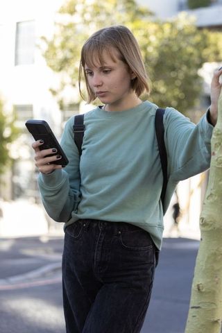 Teen girl texting on smartphone while standing on sunny tree-lined urban street with backpack