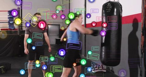 Female boxer punching heavy bag with trainer amid augmented reality social chat icons