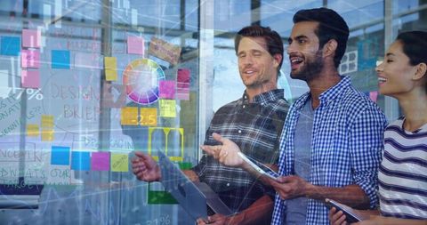 Startup team collaborating at glass wall with sticky notes, color wheel and digital devices