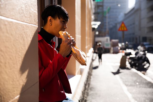 Fashionable biracial transgender person enjoying a sandwich on a city street. Suitable for use in lifestyle blogs, urban living articles, food and dining promotions, and content focusing on modern, diverse lifestyles.