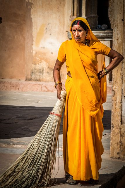 This image features an Indian woman dressed in a traditional yellow sari sweeping the streets of a village with a handmade broom. The vibrant color of her sari and the rustic surroundings highlight the cultural and traditional aspects of rural India. This image can be used for topics on Indian culture, rural life, traditional clothing, everyday activities, and gender roles in different societies.