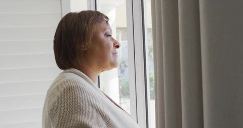 Mature woman standing by glass window looking out, cozy knit cardigan, profile silhouette