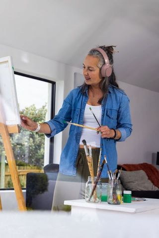 Senior woman painting at easel wearing pink headphones in sunlit home studio