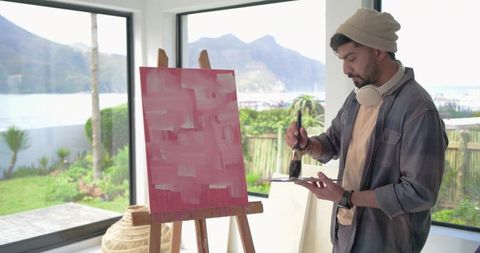 Indian man painting abstract pink canvas by sunlit window with coastal landscape view