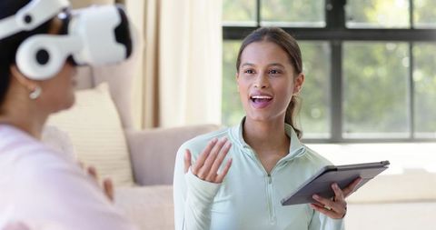Diverse female facilitator guiding vr participant with tablet in living room therapy