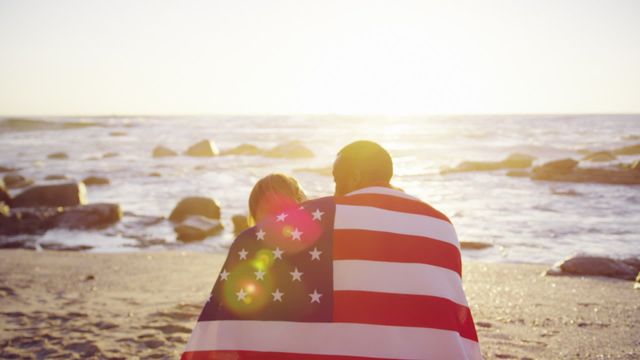 Couple sitting closely wrapped in an American flag on a beach during sunrise or sunset, signifying unity, love, and patriotism. Suitable for themes of patriotism, romance, vacations, and togetherness. Perfect for use in marketing campaigns, advertisements for beachfront resorts, patriotic events, or romantic getaways.