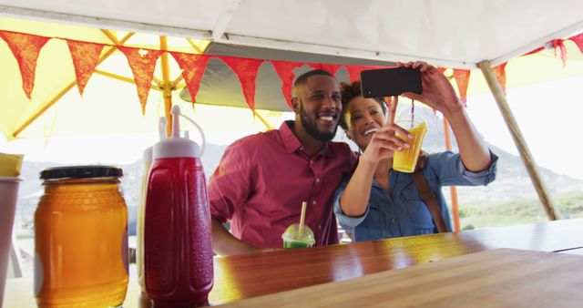 Couple taking selfie at outdoor juice bar, both smiling and holding their drinks. Bright, summery setting with cheerful atmosphere. Ideal for use in travel promotions, lifestyle blogs, outdoor event advertising, or social media content highlighting fun and leisure activities during summer.