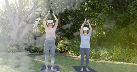 Women practicing vr yoga and stretching on sunlit lawn with virtual reality headsets