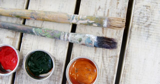 This image depicts two well-used paintbrushes and three small containers of brightly colored paints on a wooden table. The brushes show signs of extensive use, adding a rustic and artistic feel. This image is perfect for use in art-related topics, websites, or advertisements promoting creativity, craftsmanship, and artistic tools.