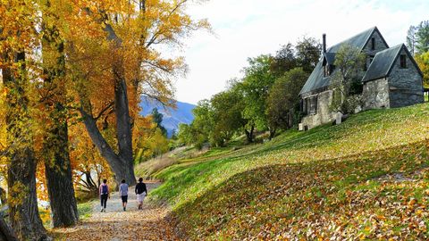 Friends Walking Autumn Lakeside Path Past Stone Cottage Under Golden Trees