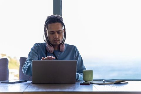African american man working remotely at ocean-view laptop with headphones and coffee