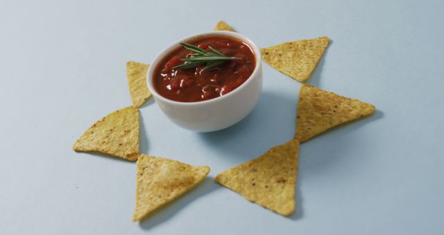 Photo showcasing a white bowl filled with spicy salsa, garnished with herbs, surrounded by arranged nacho chips. Useful for illustrating Mexican cuisine, party snacks, recipe blogs, or advertising food products.