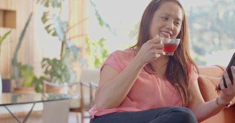 Relaxed Asian Woman Enjoying Tea while Browsing Smartphone at Home
