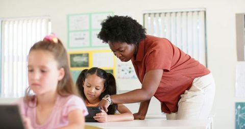 African American Teacher Assisting Elementary Student with Tablet in Bright Classroom
