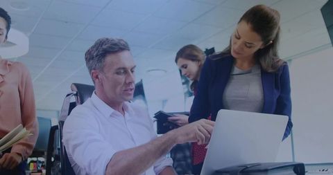 Mature manager mentoring team at laptop while colleagues collaborate in bright open office