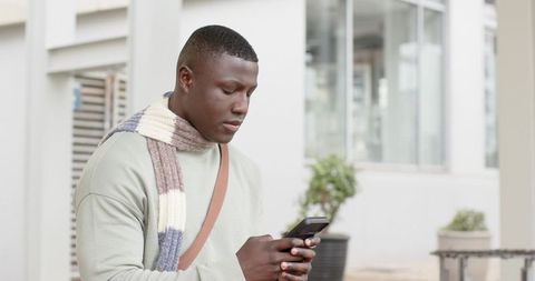 African American man checking smartphone in urban plaza wearing knit scarf and bag