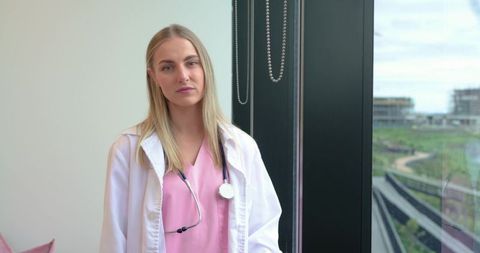 Female clinician standing by window in modern clinic wearing pink scrubs and stethoscope