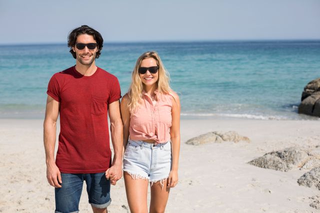 Young couple enjoying a sunny day at the beach, wearing casual summer clothing and sunglasses. Ideal for use in travel brochures, vacation advertisements, lifestyle blogs, and social media posts promoting beach destinations and summer activities.