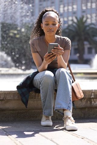 African american woman sitting on fountain edge using smartphone in sunlit urban plaza